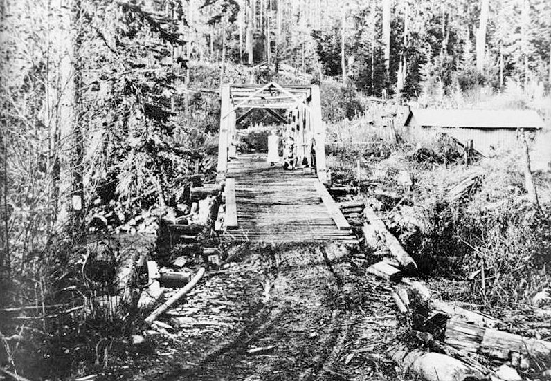 Nooksack Falls Hydroelectric Plant, Glacier Washington Historic view of timber through Howe truss bridge (replaced the earlier king post span), crossing the Nooksack River west of the powerhouse; Excelsior mine property on opposite side of river; tent frame (left), picket fence of company house, and transmission line is visible in upper left and center; looking southwest. (Photographer unknown, circa 1906).
