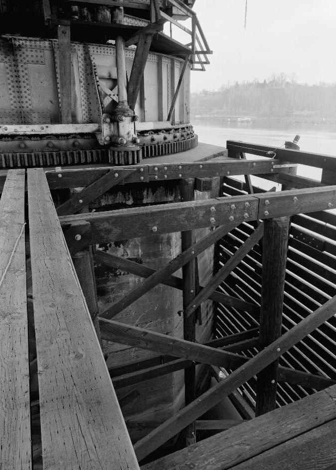 Willamette River Swing Truss Railroad Bridge, Portland Oregon 1985 BULL SHAFT, BULL RING AND PINION, AND DRUM. TOP OF PIER III, GRANITE COPING, AND PLAIN CONCRETE PIER BELOW. DETAILS OF WEST PIER PROTECTION FRAMING AT PIER. WILLBRIDGE IN BACKGROUND.